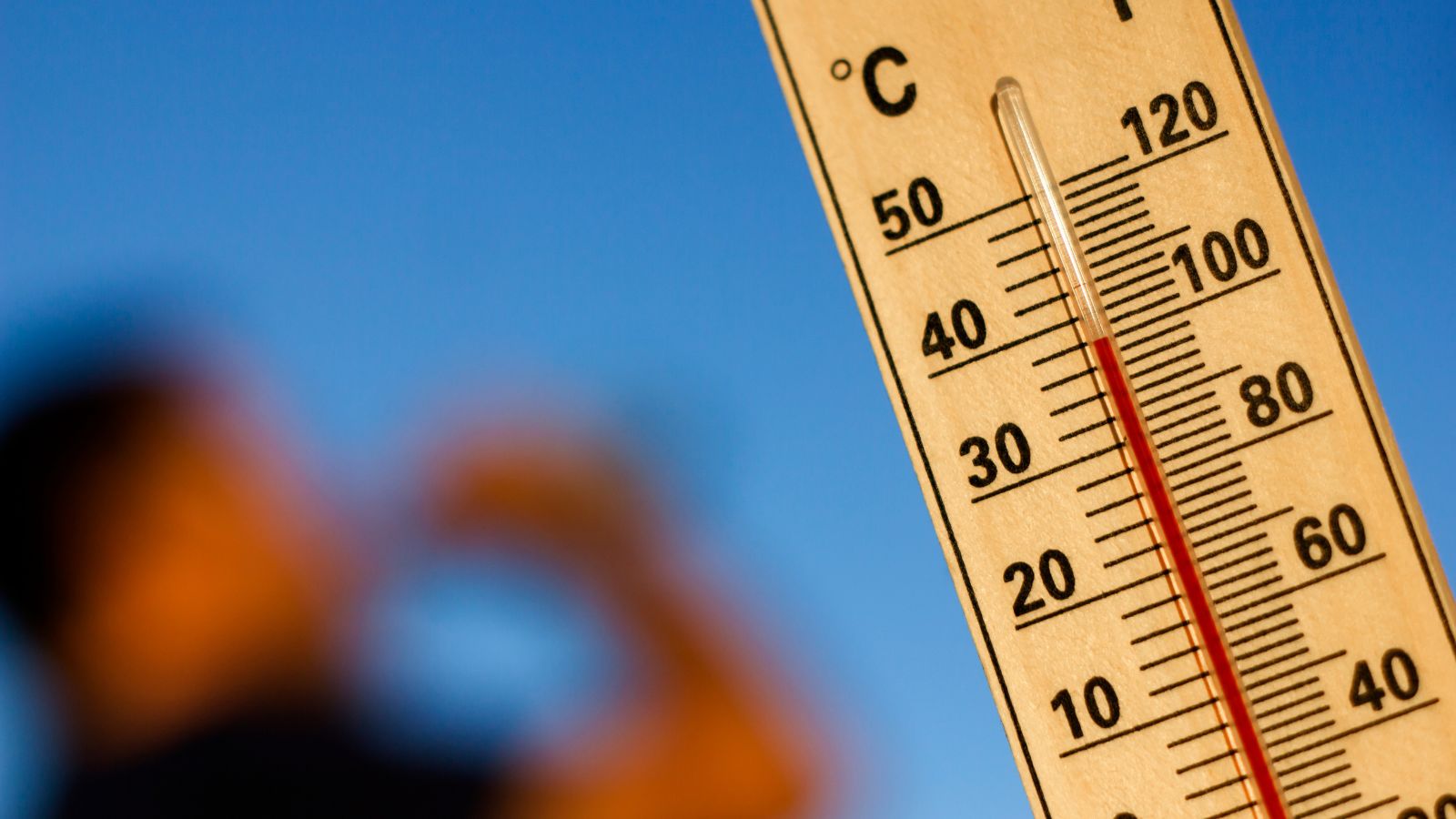 A thermometer is focused on which shows high temperatures while a man is seen in the background drinking water.